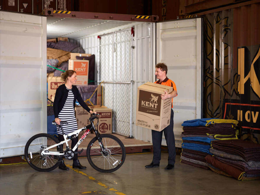 Woman holds bicycle in front of a Kent Storage container, next to a Kent Employee who holds a Kent Moving Box inside a Kent Storage warehouse.