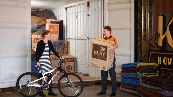 Woman holds bicycle in front of a Kent Storage container, next to a Kent Employee who holds a Kent Moving Box inside a Kent Storage warehouse.